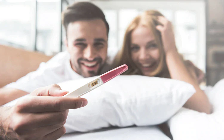 Eine Nahaufnahme eines glücklichen Paares, das im Bett liegt und einen positiven Schwangerschaftstest in der Hand hält. Die Frau lächelt strahlend und hält sich am Kopf, während der Mann stolz den Test mit zwei sichtbaren Linien in die Kamera hält. Das Bild symbolisiert die Erfüllung des Kinderwunsches und eine erfolgreiche Familienplanung, die trackle unterstützt.