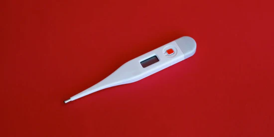 Ein digitales Basalthermometer in Weiß mit einem orangen Knopf und einem kleinen Display, flach liegend auf einem kräftigen roten Hintergrund. Das Bild zeigt ein klassisches Gerät zur Messung der Basaltemperatur im Rahmen der natürlichen Familienplanung (NFP).