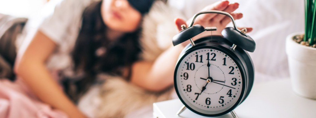 Eine Frau liegt mit einer Schlafmaske im Bett und greift mit der Hand nach einem klassischen schwarzen Wecker auf dem Nachttisch. Das Bild illustriert die Unabhängigkeit von festen Messzeiten bei der Nutzung des trackle Zykluscomputers.