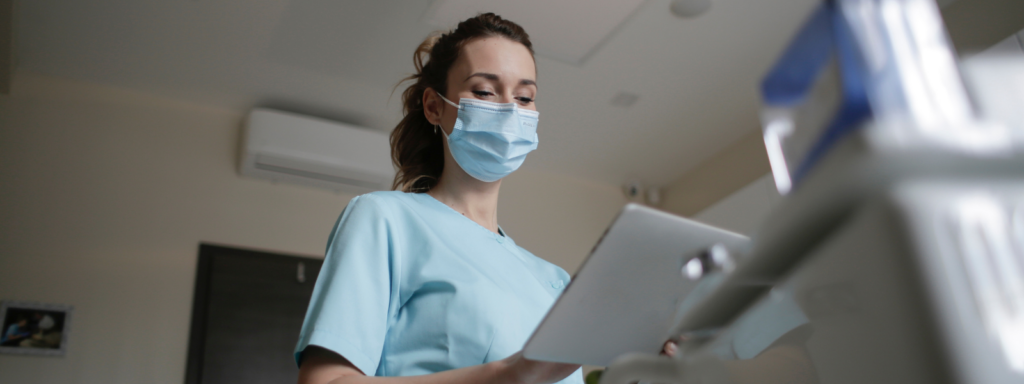 Eine Frau in medizinischer Arbeitskleidung (Kasack) und mit Mundschutz blickt konzentriert auf ein Tablet. Das Bild illustriert die digitale Zyklusauswertung mit trackle im beruflichen Kontext oder die Eignung des Sensors für medizinisches Fachpersonal mit unregelmäßigen Arbeitszeiten.