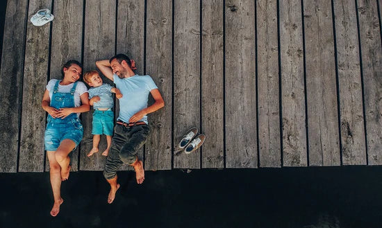 Eine Draufsicht auf eine glückliche junge Familie, die entspannt auf einem Holzsteg an einem Gewässer liegt. In der Mitte liegt ein kleiner Junge, flankiert von seinen lachenden Eltern, die sich ihm liebevoll zuwenden. Das Bild symbolisiert die Erfüllung des Kinderwunsches und ein natürliches Familienleben mit trackle.