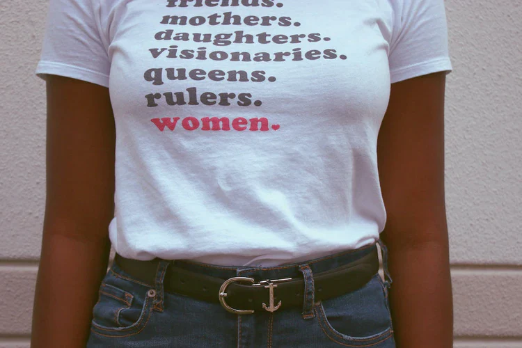 Eine Nahaufnahme einer Frau, die ein weißes T-Shirt mit einem aussagekräftigen Textaufdruck trägt: "friends. mothers. daughters. visionaries. queens. rulers. women.". Das Wort "women." ist in rot und mit einem Herzchen am Ende hervorgehoben. Das Bild symbolisiert Female Empowerment, Selbstbestimmung und die Kraft von Frauen – Werte, die trackle unterstützt.