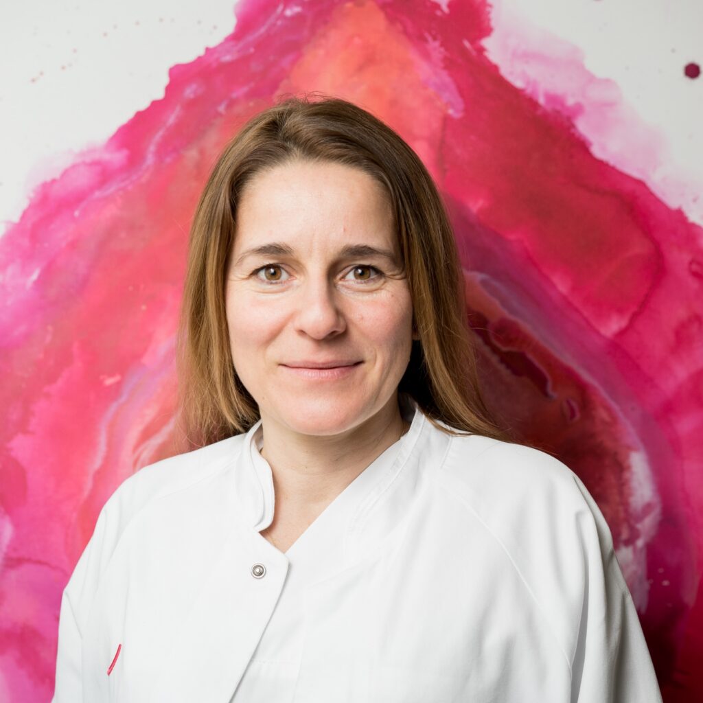 orträt von Dr. Mandy Mangler, Chefärztin für Gynäkologie und Geburtsmedizin, im weißen Arztkittel vor einem rosa-rot aquarellierten Hintergrund.