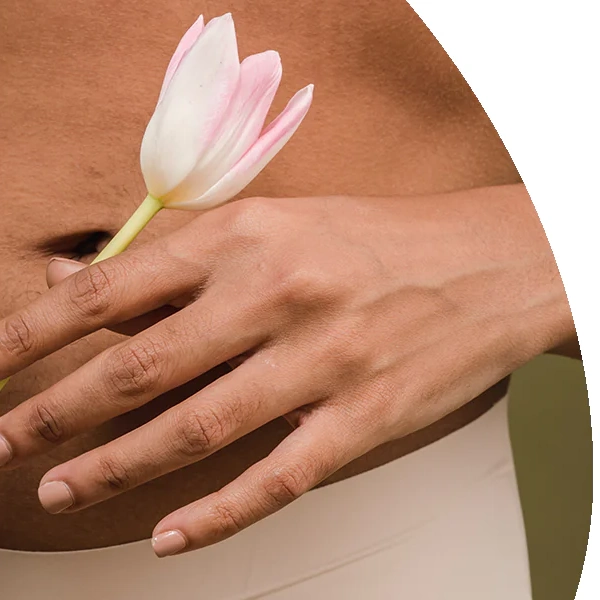 Eine Nahaufnahme einer Hand mit hellrosa lackierten Nägeln, die sanft auf einem Bauch ruht. Die Finger umschließen locker den Stängel einer einzelnen, weiß-rosa Tulpe. Die Haut ist im warmen Ton gehalten, der Hintergrund ist neutral. Das Bild symbolisiert Weiblichkeit, Achtsamkeit gegenüber dem eigenen Körper und die Verbindung zum Zyklus.
