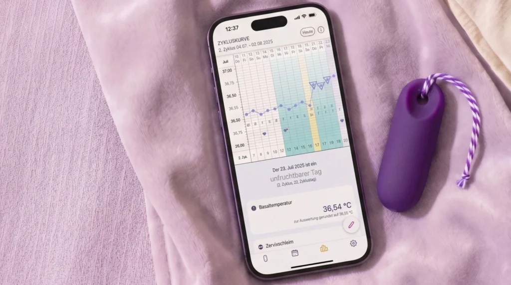 Ein Smartphone liegt neben dem kleinen lila trackle Sensor auf einer hellvioletten Decke. Auf dem Bildschirm ist eine Zyklus-App mit einer Kurve zur Basaltemperatur und dem Text „unfruchtbarer Tag“ auf Deutsch zu sehen.