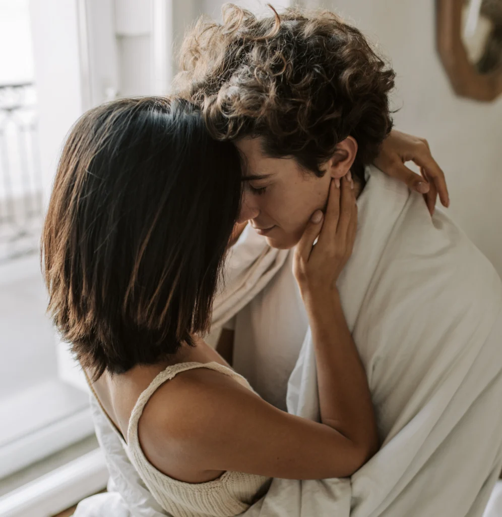 Ein junges Paar in einer innigen Umarmung im Schlafzimmer. Die Frau hat kurze, dunkle Haare, der Mann lockiges Haar und ist in eine weiße Decke gehüllt. Sie berühren sanft ihre Gesichter.