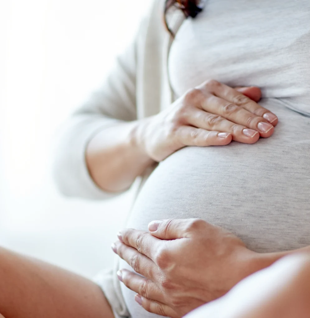 Nahaufnahme einer schwangeren Frau in heller Kleidung, die behutsam beide Hände auf ihren runden Babybauch legt.