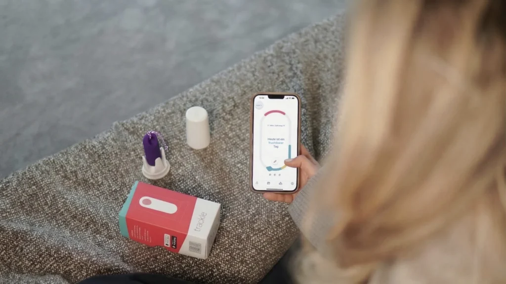 Eine Ansicht über die Schulter einer Frau, die ihr Smartphone hält. Auf dem Display der trackle App ist die Meldung „Heute ist ein fruchtbarer Tag“ zu sehen. Neben ihr auf einer strukturierten Oberfläche liegen der lila trackle Sensor in seiner Halterung sowie die Produktverpackung. Das Bild verdeutlicht die digitale Zyklusauswertung in der täglichen Praxis.